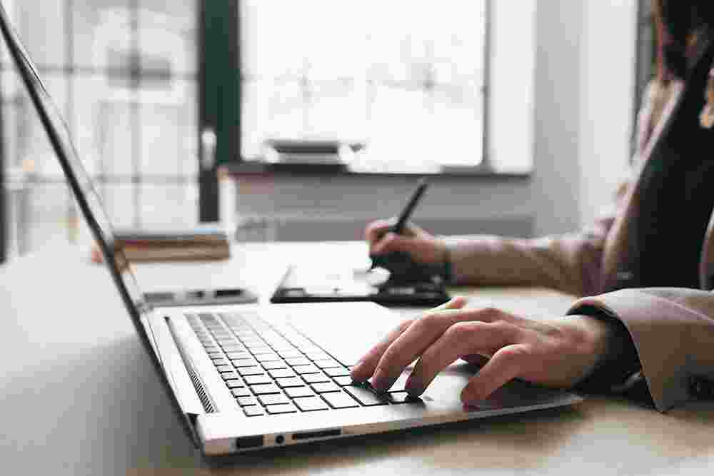 Woman holds one hand on the laptop while writing something down.